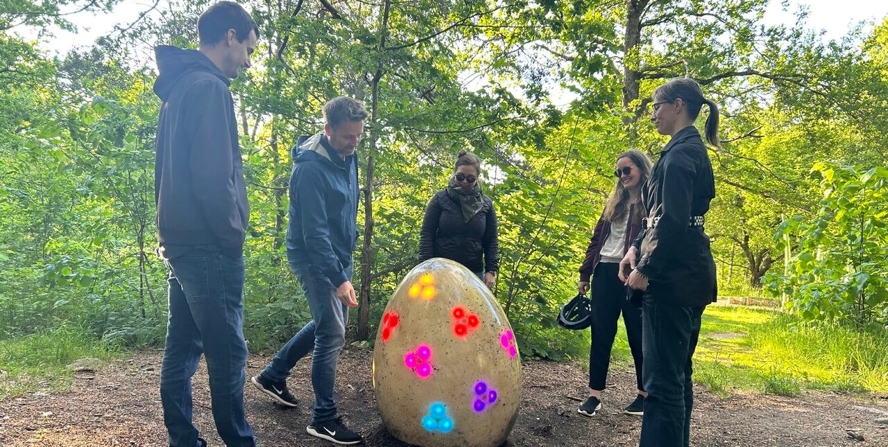 Drakryggens team står runt en stor ägg med färgglada lysande ljus, omgivna av grönskande träd i Magistratshagen lekpark.